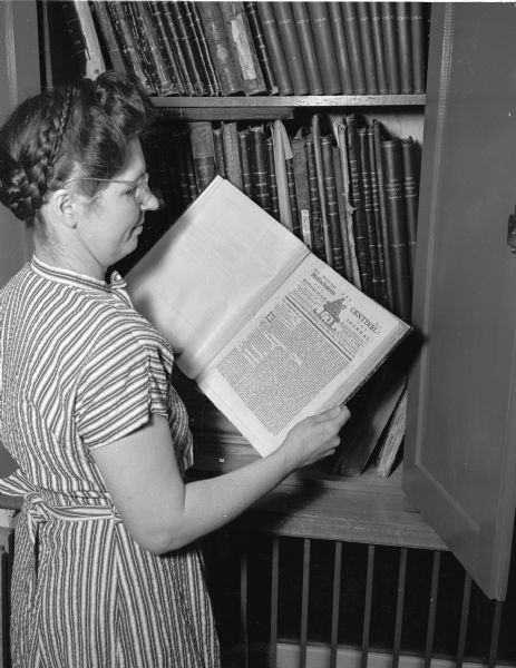 Mrs. Robert (?) Nelson, librarian, at the Wisconsin State Historical Library, standing before a case containing a rare collection, and looking at a copy of the July 7, 1784, <i>Massachusetts Centinel</i>, published in Boston.