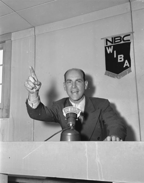 Arthur Bramhall, at a microphone, gesturing as he announces a sports event. Behind him is the identifying emblem for NBC WIBA radio affiliate.