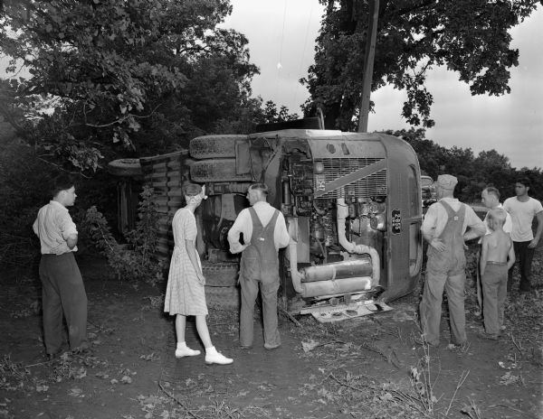 Several bystanders are observing the damage to a Greyhound bus turned over on its side as the result of an accident on Highway 12 about 2 miles east of Madison.  The driver and forty-one passengers were injured but all survived when the bus careened through the air, smashing trees, fences and a cement culvert.