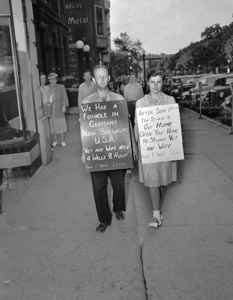 Mr. and Mrs. Sidney Levy, wearing sandwich boards stating "We Had a Foxhole in Germany, Now Sidewalks, USA. Vet and Wife Need 4 Walls & Roof" and "After September 1st, The Street Is Our Home. Open Your Home to Student Vet and Wife." The couple is walking on the Capitol Square.