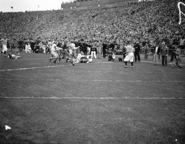 Fullback Earl Maves (35) running off tackle for eight yards to the Ohio State one yard line accompanied by University of Wisconsin-Madison end Dale Bowers (93). He was downed by two Ohio State Players, one of whom was halfback Tommy James (66). A University of Wisconsin male cheerleader looks on.
