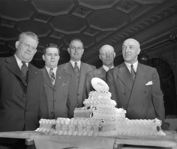 Officers of the East Side Business Men's Association posing with the tier cake made for the Association's Silver Jubilee banquet at the Hotel Loraine. Pictured from the left are: Alf Kjeverud; Trygve Strand; Ray Sennett; Lars Landsness; and L.L. Lunenschloss, banquet host.