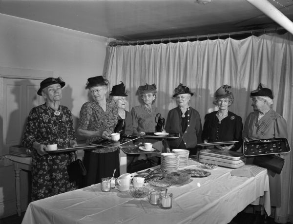Seven ladies of the Women's Guild of Pilgrim Congregational Church, 953 Jenifer Street, holding trays and standing around a reception table, observing the fifty second anniversary of the founding of the guild.  Left to right: Mrs. Marilla Ward, Mrs. S.A. Cripps, Mrs. H.O. (Amy) Bigelow, Mrs. Effie Appleby, Mrs. Bessie Goodman, Mrs Eugene (Mabel) Lemon, and Mrs. John (Helen) Ralph. Mrs. Appleby was the founder of the guild in 1895.