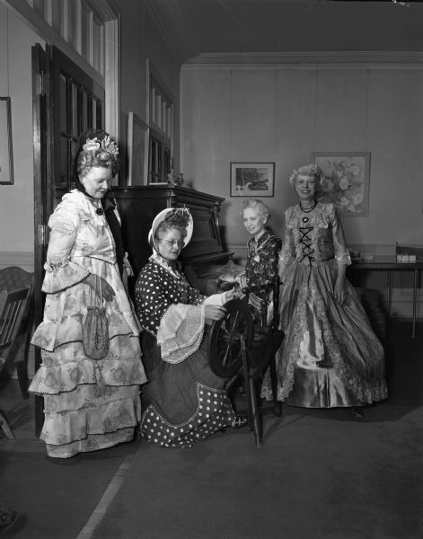 Four members of the Who's New Club, in Civil War costumes. One woman is seated at a piano, one sitting near a spinning wheel, and two standing. They are participating in the 12th anniversary of the club.  Left to right: Committee members, Mrs Carson A. Hatfield, Mrs. A.F. (Laura) Toepelman, Mrs. (Ruth) Arthur L. Luedke, and Mrs. T.M. (Ida) Sprowls.