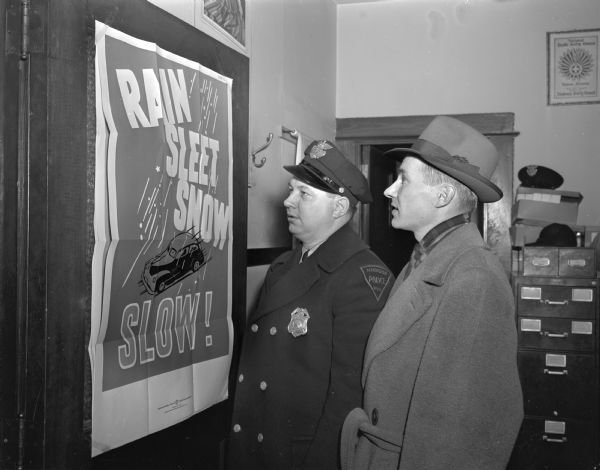 Observing a poster stating: "Rain, Sleet, Snow, slow!," are Police Officer Walter Randall, with Edwin B. Petersen, winner of the "Wisconsin State Journal's" courteous driver campaign.