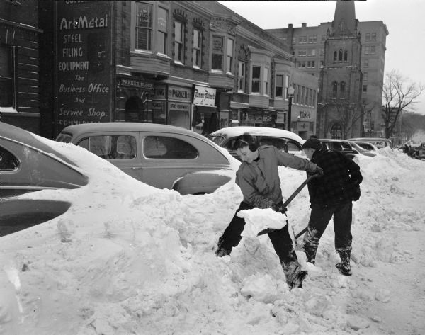 Winter scene with Dave Brown (left), and Bill Pedder shoveling out cars parked on South Carroll Street on the Capitol Square, charging $1.50 and up.