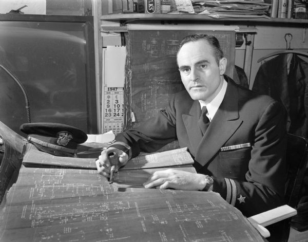 Gordon E. Harmon, Superintendant of Construction for George Nelson and Sons, General Contractors, shown in uniform at his desk surrounded by blueprints. He is commanding officer of the Ninth Naval District's reserve unit in Madison.