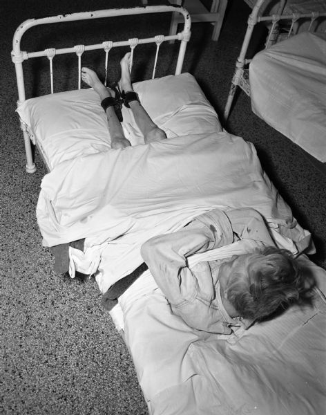 A woman patient at the Mendota State Hospital (Mendota Mental Health Institute) who is restrained in her bed by a camisole, which prevents the use of her arms, while her feet are strapped to the foot of her bed. One of a series of photographs calling attention to the inadequacies in personnel and facilities at the hospital.