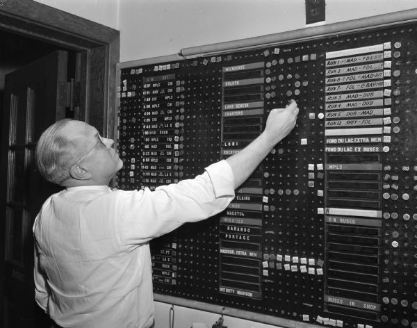 Frank Sager, chief dispatcher of the Northland Greyhound Lines in Madison, shown at a board listing the names of the drivers, the buses they are driving, and their location around the state.