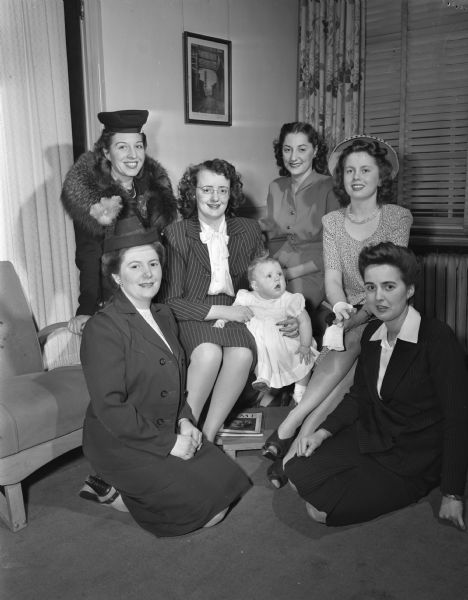 Group portrait of six war brides and one infant. Nine-month-old Susan Schreiber can claim both American and Australian citizenship. Shown playing with the baby are Mrs. Franklin P. Hopkins, president of the new Overseas club, kneeling at the left, and left to right: Mrs. Raymond Hovde; Mrs. Alvin Schreiber; Mrs. Irwin Bosman; Mrs. M.B. Wheeler; and Mrs. Kenneth Kitchen. Mrs. Kitchen comes from London; the rest are natives of Australia.