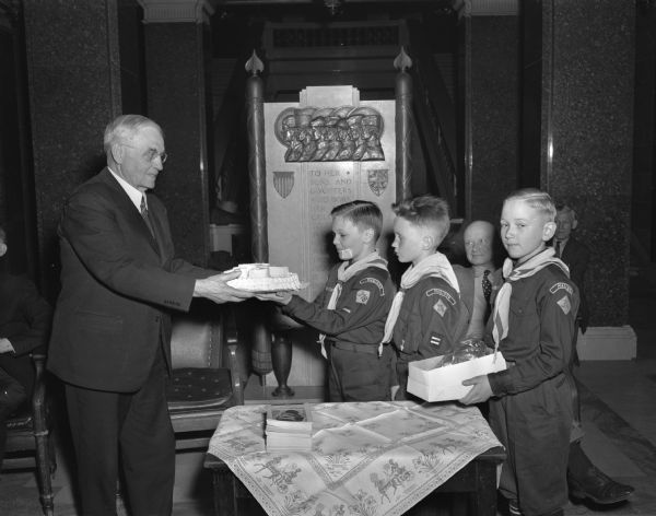 Because his birthday falls on the same date as Abraham Lincoln's, Chief Justice Marvin B. Bosenberry, honorary president for the Four Lakes Boy Scout Council, was presented with a birthday cake in honor of his 79th birthday by three representatives of Cub Pack No. 318. This was part of Lincoln's Birthday observance held in the Wisconsin State Capitol Wednesday.  From left to right are: Bernard Coughlin, John Feeny, and David Johnson.