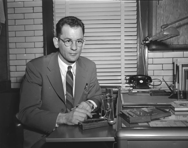 Malcolm W. Jensen, age 30, City of Madison Sealer of Weights and Measures, is pictured at his desk in the Madison City Market Building.  The objects on his desk are a metric weight and a one-once balance used for comparison and calibration of pharmacists' prescription weights and jewelers' weights.  He encouraged the city to buy a Toledo automatic dial-type truck scale available to both the city and commercial users.