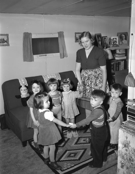 Mrs. Milton Sager, in a trailer home at Camp Randall, does day care with six children. The trailer park, provided by the University of Wisconsin, is for WWII veterans and their families. Children in the day care are: (left to right) Carol Sager, Linda Jean Scheldrup, Ellen  Cartier, Carol Jacobson, Peter Mattila, and Jimmy Cartier.