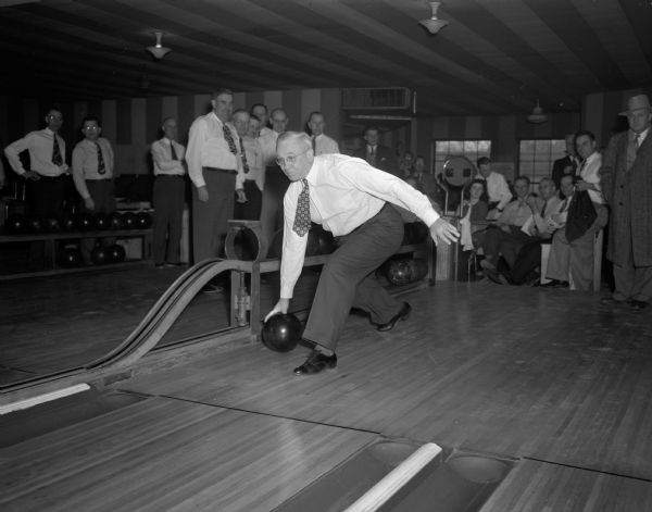 A bowler is delivering his ball at the Kiwanis State Bowling Tournament held at Schwoeglers, the Plaza, and Moderne Recreation, watched by other bowlers.
