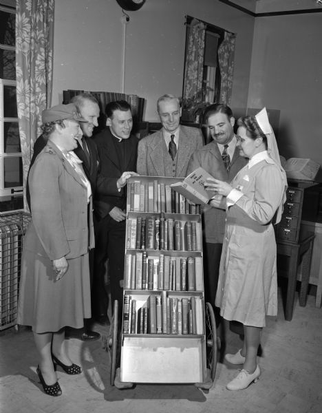 The Red Cross Gray Ladies continue to offer their services at Wisconsin Memorial Hospital for veterans on the grounds of Mendota State Hospital.  Two Gray Ladies are shown in the library with four hospital officials. Left to right:  Shyrle Frederickson Nelson, Fred M. Ritzman, special services, Rev. Edward B. Auchter, chaplain, Rev. James R. Love, chaplain, Louis Fishbune, medical rehabilitation, and Mrs. Gordon (Eva) Adams.