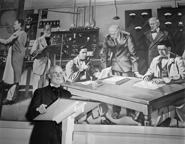 Dr. William H. Lighty, one of the guiding forces behind Radio Station WHA in its infancy, is shown at the lower left, duplicating the position he was in back in the 1920's in the mural behind him. The mural is of early day operation of the station. Seated at the table in the mural is Malcolm Hanson. Standing next to him is Prof. Andrew Hopkins, with Prof. Edwin Bennett standing at the right. Seated at the right is Prof. E.M. Terry.