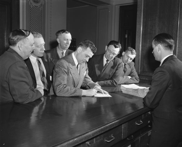 Glenn Davis signing his nomination papers for Gaige S. Roberts in the Secretary of State's office. Davis was filing to become a Republican candidate for Congress. Supporters with him are, left to right, Senator Frank Panzer, Oakfield; Assemblyman David Dancey, Waukesha; Alfred Ludvigsen, Hartland; Elmer Genzmer, Mayville; and Arnie Betts, Lodi.
