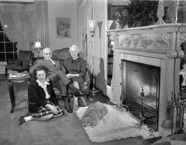 Group portrait of Governor Oscar Rennebohm and his family in the living room of their home at 201 Farwell Drive, Maple Bluff. Pictured in front of the fireplace left to right: daughter Carol, the governor, and Mrs. Rennebohm (Mary) and their dog.