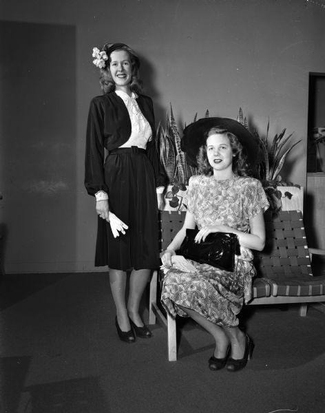 University of Wisconsin Women's Self Government Association style show, showing two women, one standing in a suit with hat and gloves, and one seated, wearing a dress and a large hat and holding a purse. Both women are wearing open-toed pumps.