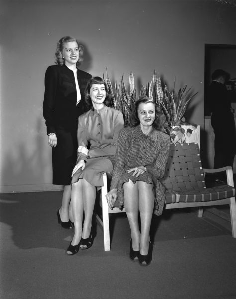 University of Wisconsin Women's Self Government Association style show "Confetti of Fashion" committee members watching a rehearsal. Jean Kerth, standing, chairman of the models, and two models seated, Jane Walker, Wauwatosa, and Betty Einhorn, Rockford, Illinois. All three women are wearing dresses with open-toed pumps.