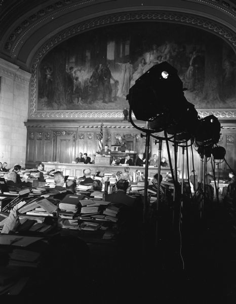 Wisconsin Assembly in the Wisconsin State Capitol being filmed for State Centennial Film. There are a group of men on the speakers rostrum, and many cameras and multiple lights for the filming of the legislators are in the foreground.