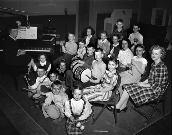 Fanny (Mrs. William) Steve, at the piano, and children playing instruments in WHA radio studio. Mrs. Steve was part of the program staff of the School of the Air, WHA.  The children are from Oak Hall School in the town of Fitchberg and the teacher is Mrs. Anderson.