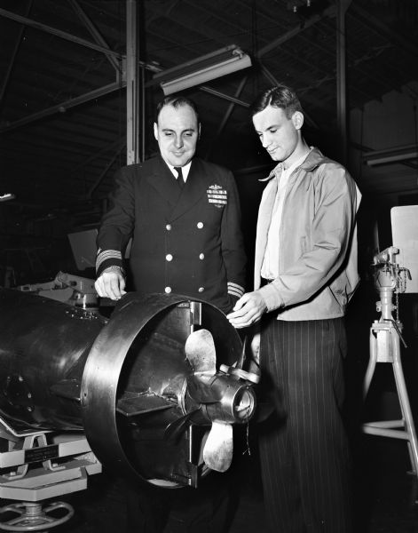 Celebrating the 47th anniversary of the purchase of the first "submersible torpedo boat" from its inventor, J.B. Holland, for service in World War I, are Commander J.C. Martin and Donald Neushown. They are examining a torpedo.