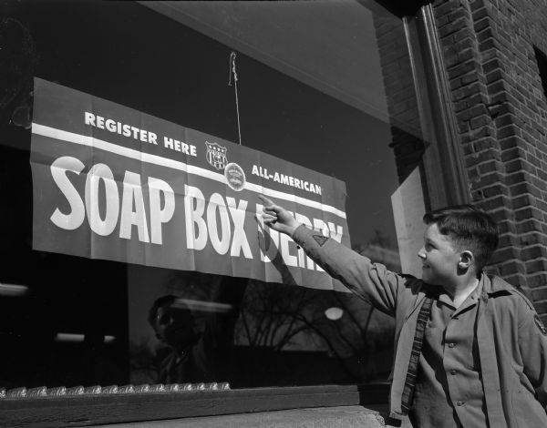 Micheal "Mickey" Lathers, son of Mr. and Mrs. William Lathers, Jr. (Gladys), 216 Virgina Terrace. Micheal is pointing to a sign in a window, "Register Here - All American Soap Box Derby."