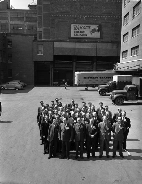 Group portrait of over 25 Oscar Mayer salesmen from Chicago shown in lot in front of Madison Oscar Mayer Plant with trucks, loading docks and billboard stating: "Welcome Chicago Salesmen".