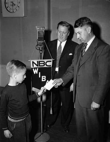 Two men presenting a bicycle safety award to a young boy in front of a NBC WIBA radio microphone.