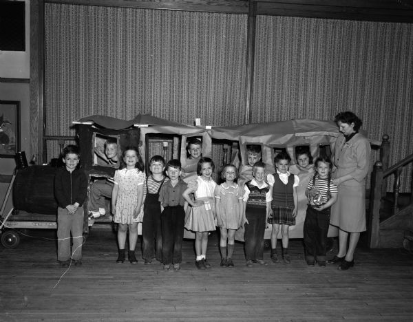 Thirteen students of the Shorewood Hills kindergarten class with their teacher posed in front of a train, constructed of paper and a large barrel, in the school gymnasium.