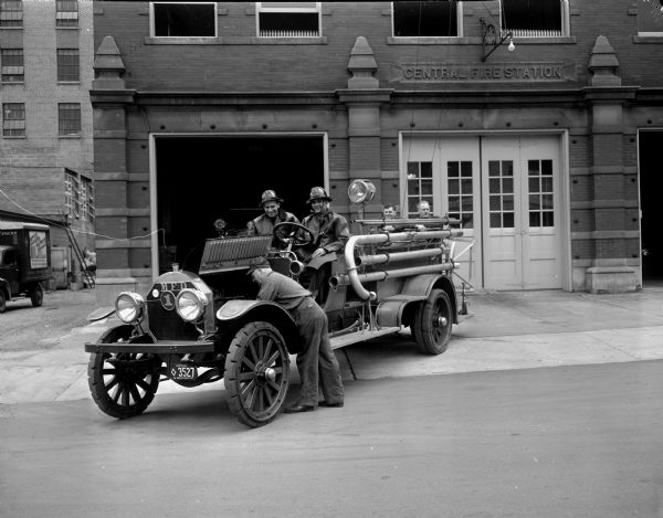 Firemen James Fraser and Philip Statz (left to right in the driver's seat) are demonstrating how the old wooden spoke-wheeled 1915 foam fire truck chugged a coughing gasp and refused to move from the Central Fire Station, 18 South Webster Street. Capt. Arne Lerwick, the master mechanic, is shown working on the motor. At the back of the fire truck (l to r) Firemen Paul Welsch and Chris Anderson.