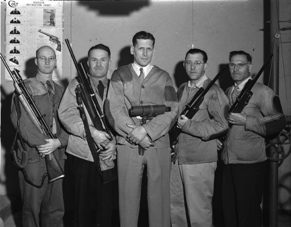 Group portrait of five men in hunting clothing, holding rifles. There is a revolver instruction chart on the wall in back of the men.