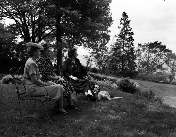 Outdoor portrait of three members of the John Bell Daughters of the American Revolution (DAR) Chapter. They are (from left to right), Edna Pike and Mildred Hanks, co-chairs of garden tour committee, and Ruth Neff in whose garden at 731 Farwell Drive, Maple Bluff, they are seated. A dog is sitting on the grass.