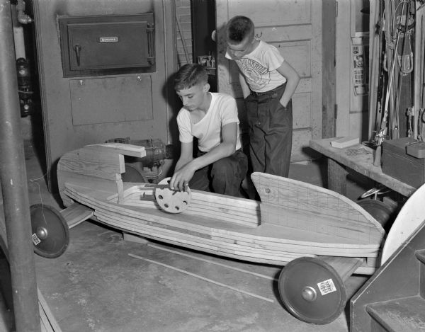 Ed Gesteland, who is fourteen years old, is shown working on his Soap Box derby racer in the basement of his home at 4221 Wanetah Trail. Behind him, with hands in pockets, stands his brother, Ray. The boys are sons of Mr. and Mrs. E.R. Gesteland.