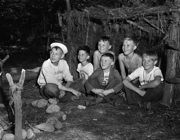 Six boys enjoying their grass covered lean-to in front of a campfire at YMCA Camp Wakanda. L to R in the foreground are: John Omernik, John Stephens, John Theis. L to R in the background are: John Wyngaard, Anthony Purnell, Peter Cash.