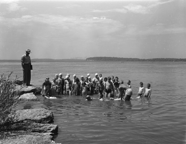Pictured at Tenney park are Sun Prairie children receiving swimming instruction from Mrs. Mildred Schwieten, standing in the water at the left, and Don Heimlich, on the right. At the extreme left standing on the rock is Francis Sheehan, water safety director of the Sun Prairie Red Cross Chapter. The Red Cross is providing swimming classes for rural children.