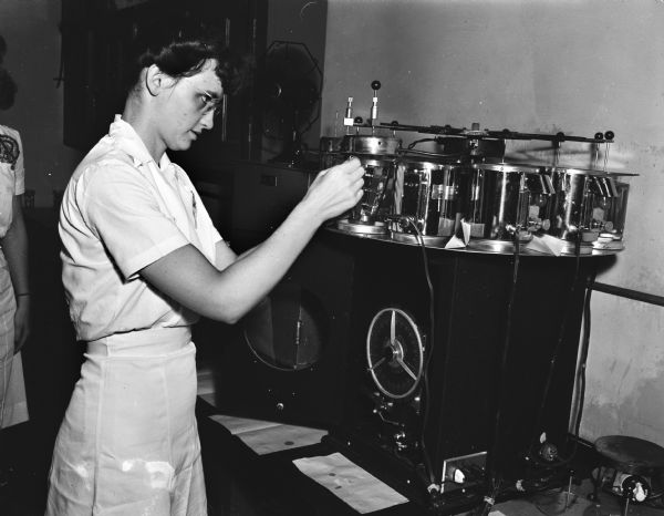 Dr. Ann Laird, resident in pathology at Wisconsin General Hospital, adjusts the technocon which speeds cancer diagnosis, in the laboratory at the Wisconsin State Laboratory of Hygiene. The round machine advances the suspected tissue from its solution in one glass jar to the next until it is ready to be made into slides for diagnosis.