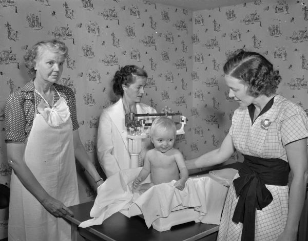 Visiting Nurse Service, sponsored by Attic Angels, at a child health center at Emerson School. This center is one of five child health centers sponsored by Attic Angels. Catherine Bassett is weighing Jill Ann Warner with Attic Angel probationer June McLean looking on. In the center is Dr. Sally Cornell Mendenhall, also a probationer, and doctor in attendance.