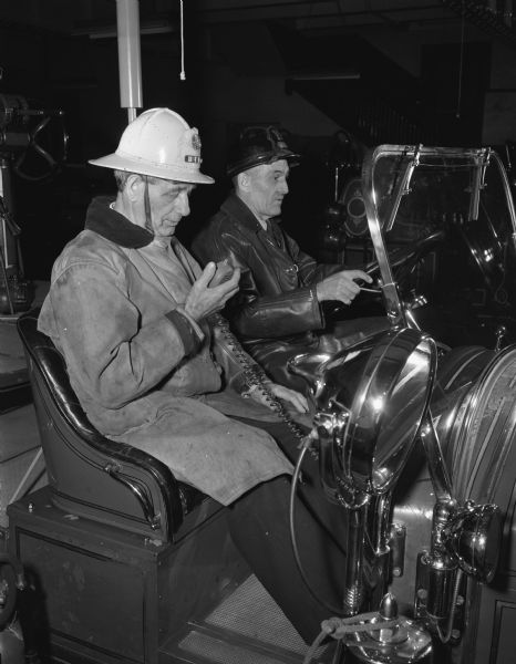 First Assistant Fire Chief Patrick J. Brown with microphone, and Fireman Grant Prideau receiving radio instructions from Fire Chief Edward J. Page, demonstrating the new mobile radio equipment. The firemen in uniform are seated in a fire truck.