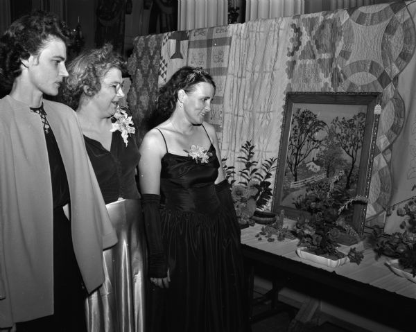 Three women in formal wear at the Hotel Loraine viewing floral arrangements set in a background of crocheted tablecloths, quilts, and embroidered cloths which were all created by members of the Sunset Village community. Pictured from left to right are; Mrs. C.E. Fagan, Mrs. T.F. Wisniewski, and Mrs. E.H. Fisher.