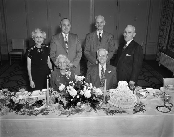 Frank & Alma Sager seated at a table with flowers, cake, and candles. Three men and one woman are standing behind them.