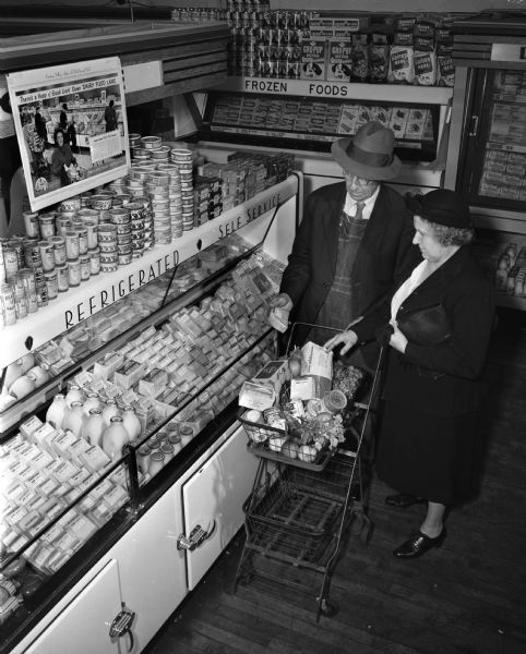 Elevated view of man and woman with a grocery cart shopping at the dairy case in a grocery store. Sign over case reads: "There's a Heap of Good Livin' Down Dairy Food Lane". Photograph taken for the American Dairy Association of Wisconsin.