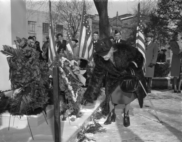 Armistice Day ceremony with Mrs. Ella M. Bresee, representing the Grand Army of the Republic and the Daughters of 1812, shown placing a wreath at the cenotaph with veterans in uniform present. The ceremony is taking place at the Wisconsin State Capitol grounds.
