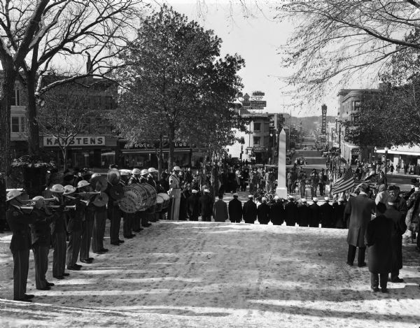 Armistice Day ceremony, looking down State Street, with a drum and bugle corps on the left and other veterans in uniform standing at attention near the cenotaph. The ceremony is taking place at the Wisconsin State Capitol grounds.
