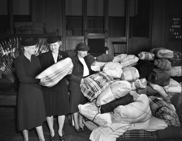 Members of the Madison Council of Church Women are shown with bundles of warm clothing and blankets, shoes and school supplies from Madison Sunday school classes and women's societies of all churches, to be sent to aid the needy people in Europe. Left to right are Mrs. Ralph A. McCanse, program chairman; Mrs. Ron L. Blodgett, president of the Madison Council of Church Women; and Mrs. Herbert Loucks, chairman of the bundle project.