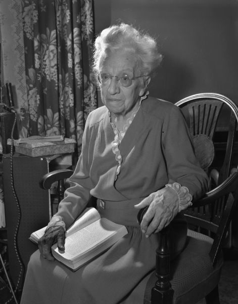Portrait of Myra (Mrs. C.F.) Cary seated in a chair holding a book in her suite at the Park Hotel on her 86th birthday. She and her husband led the way in making Wisconsin a pioneer in education for exceptional children.