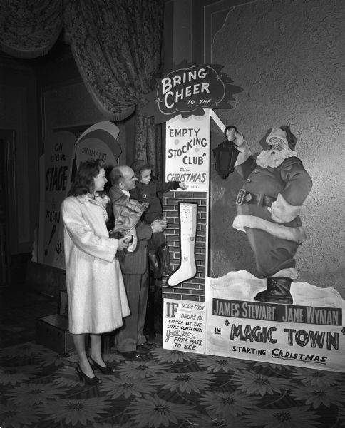 In the lobby of the Capitol Theatre, manager Fred Reeths is holding Johnny Heitz while he drops a coin into the Empty Stocking Club stocking. Johnny's mother, Virginia Heitz, looks on.  A large poster of Santa Claus is in the background.