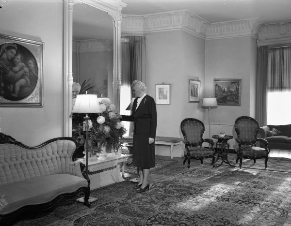 Mrs. Oscar (Mary) Rennebohm arranging flowers in the drawing room of the Wisconsin Governor's residence, 130 East Gilman Street. The house is 93 years old and has high ceilings in the rooms, and Victorian furnishings.