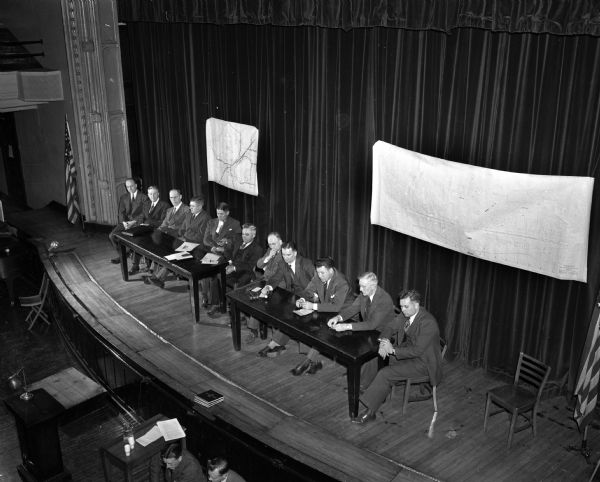 Elevated view of eleven men sitting on the stage at Central High School auditorium conducting a hearing on the possibility of putting West Washington Avenue under the Chicago, Milwaukee, St. Paul and Pacific Railroad tracks.